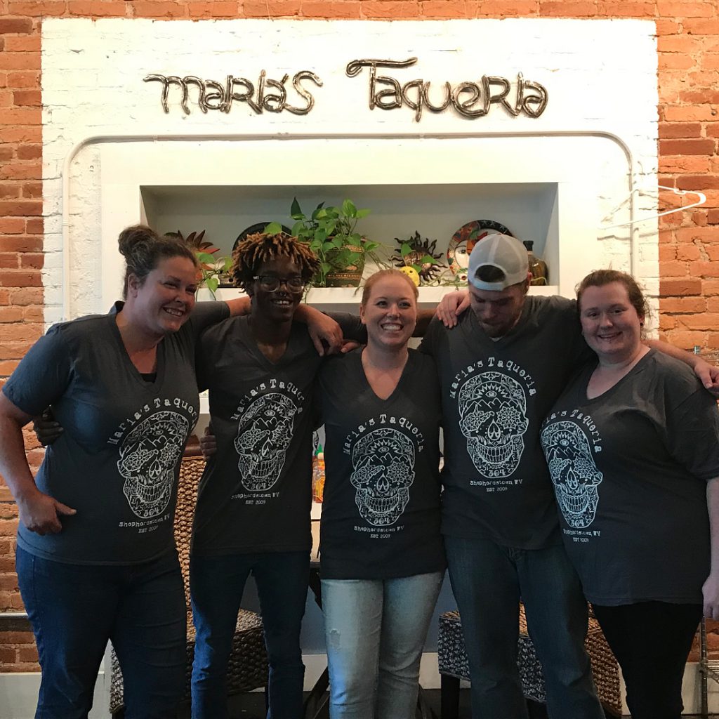 Group of five smiling staff members wearing matching gray t-shirts featuring a skull design, standing in front of a decorative wall with the text "Maria's Taqueria," showcasing a vibrant and welcoming atmosphere for a restaurant setting.