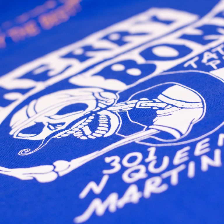 Blue t-shirt featuring a white graphic of a skull with crossed guns and the text "Cherry Bomb Tattoo" and "301 N Queen Martin."
