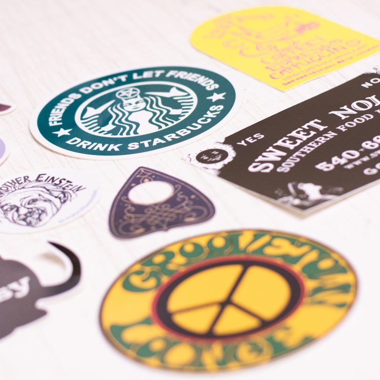 Variety of colorful custom stickers featuring logos and designs, including "Friends Don't Let Friends Drink Starbucks," "Sweet Nola's Southern Food," and a peace symbol, representing Hau Ink's printing services.