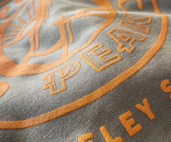 Close-up of a gray fabric featuring orange embroidered logo text that reads "PEAK" and additional branding details, highlighting custom apparel design relevant to Hau Ink's offerings.